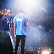 Two people on stage with a crowd, one in a blue shirt with 'Teck' on the back, light shining down.